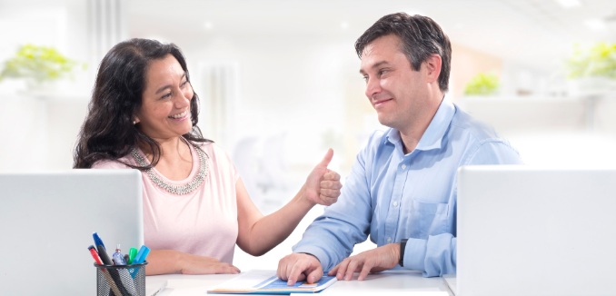 Compañeros de trabajo mirándose, sonriendo y la mujer tiene una mano levantada haciendo la señal de “bien”